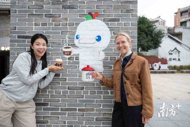 來自英國的游客（右）手持十五運特色杯子與喝咖啡的“喜洋洋”墻繪合影。